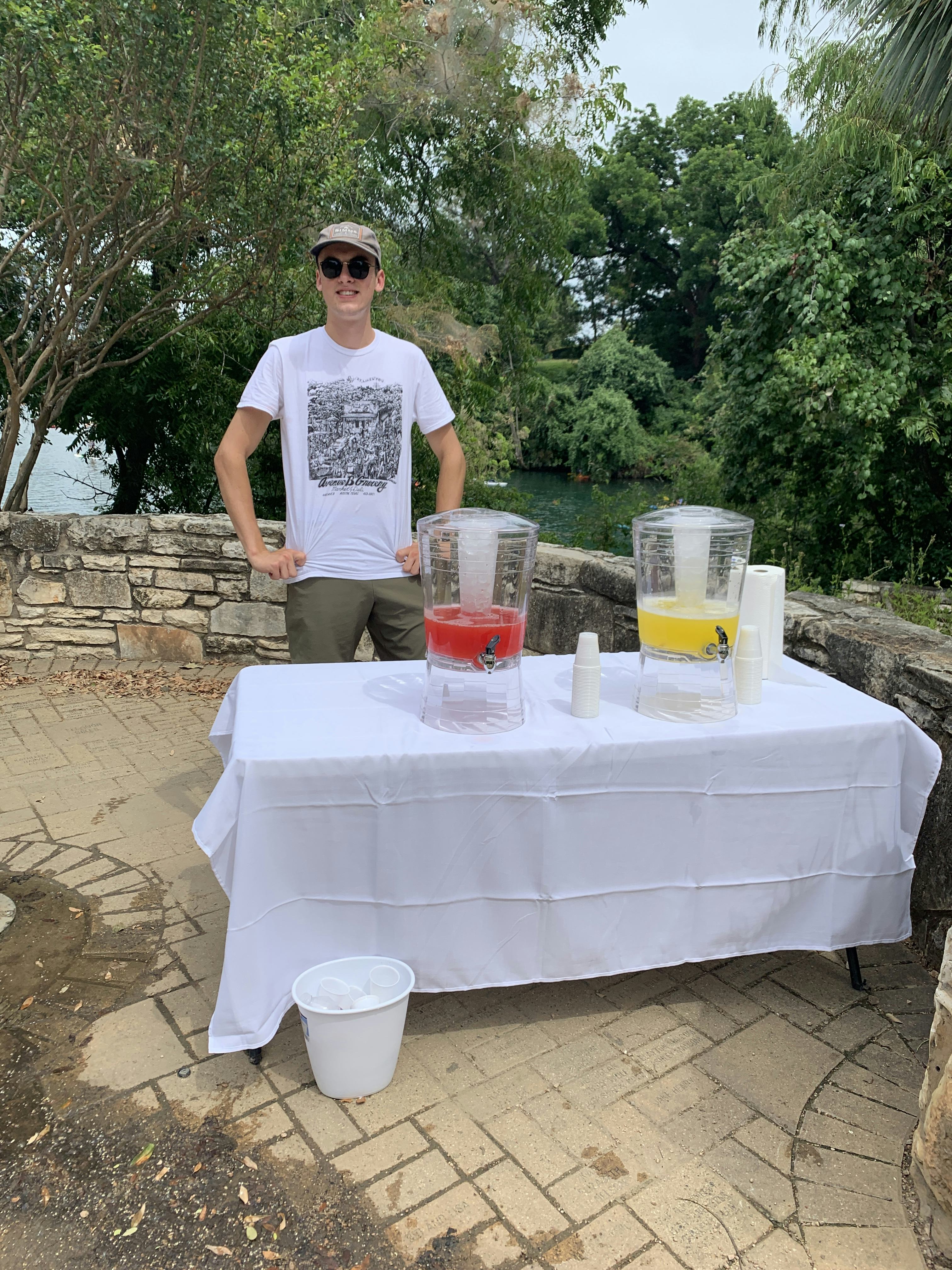 Jordan Hicks behind a table with agua fresca beverage dispensers at the Farmer's Market